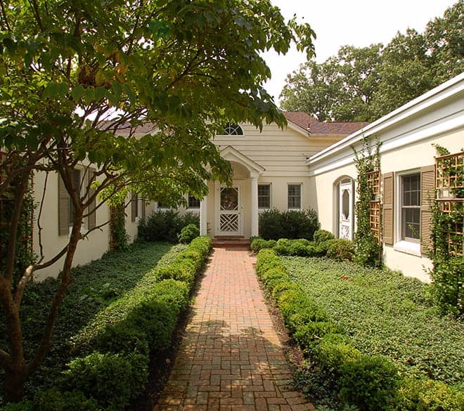 Landscaped brick walkway leading to a traditional home surrounded by mature trees, shrubs, and garden greenery.
